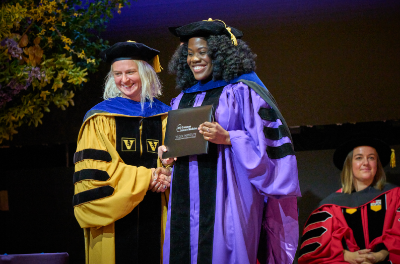 Two NYU Vilcek Institute faculty members shaking hands with a graduate holding her diploma at commencement